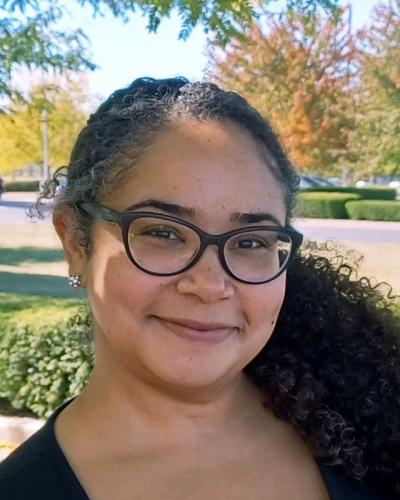 A woman with curly hair and glasses smiles at the camera in front of a park-like setting.