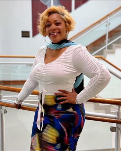 A woman with short blonde hair smiles while posing with one hand on her hip and the other on a railing.