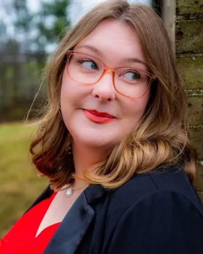 A woman with shoulder-length hair and orange-rimmed glasses smiles at the camera while leaning against a brick wall outdoors.