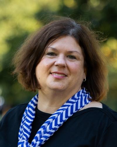 A woman with brown hair and a blue and white patterned scarf smiles while looking off to the side in an outdoor setting.