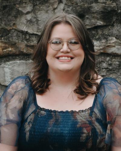 A woman with brown hair and glasses smiles while standing in front of a stone wall.