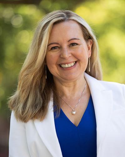 A woman with blonde hair and a warm smile, wearing a white blazer and blue shirt, stands in front of a blurred outdoor background.