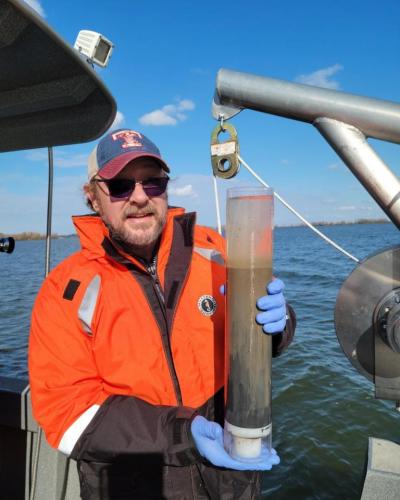 A man in an orange jacket holds a cylindrical container filled with brown liquid on a boat over water.