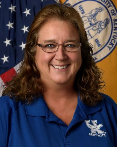 A smiling woman with shoulder-length hair and glasses wearing a University of Kentucky Army ROTC polo shirt in front of an American flag and University of Kentucky flag.