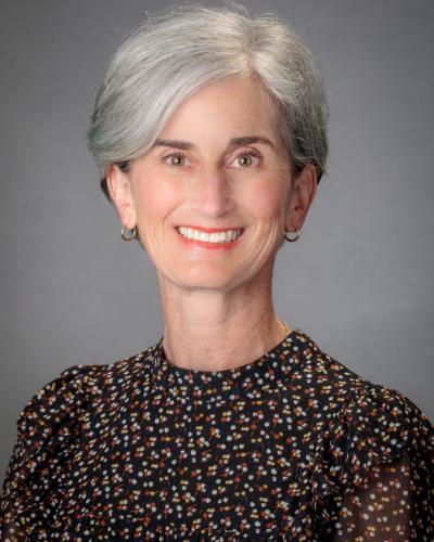 A woman smiles while looking off to the right, wearing a dark blouse with a small colorful pattern and gold earrings.