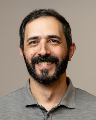 A man with dark hair and a beard smiles at the camera wearing a gray polo shirt.