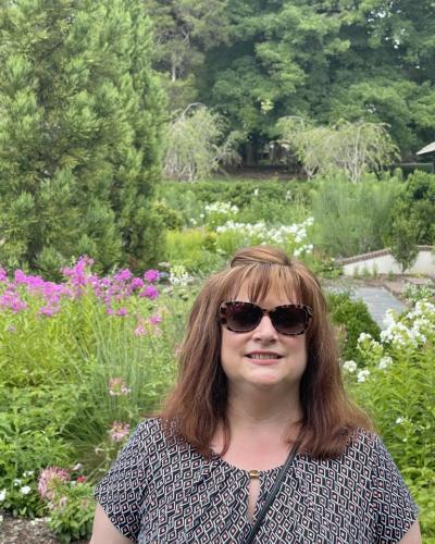 A woman stands in front of a garden filled with a variety of flowers and trees.