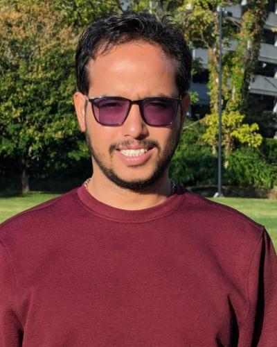 A man wearing sunglasses and a maroon shirt smiles while standing in front of a park.