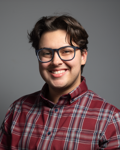 A man wearing a red plaid shirt and blue-framed glasses smiles at the camera against a gray background.