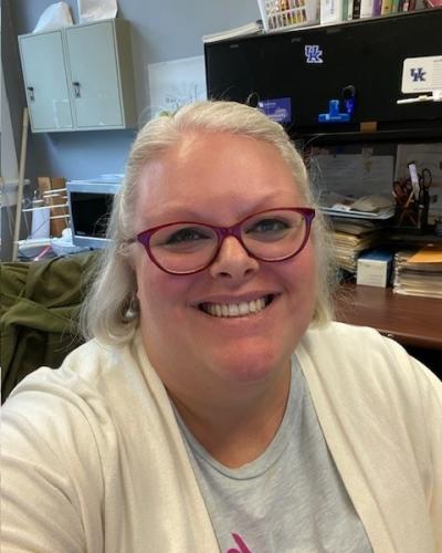 A woman with blonde hair and glasses smiles while sitting at a desk in an office.