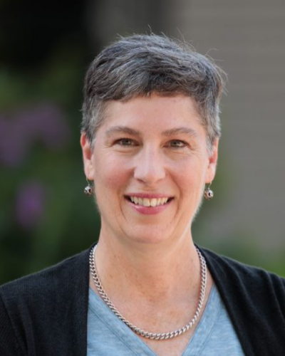 A smiling woman with short hair and a silver necklace and earrings wearing a blue shirt and black cardigan.
