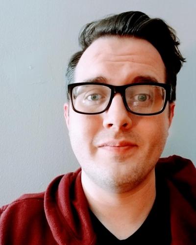 A man wearing black-frammed glasses and a red hoodie looks at the camera.
