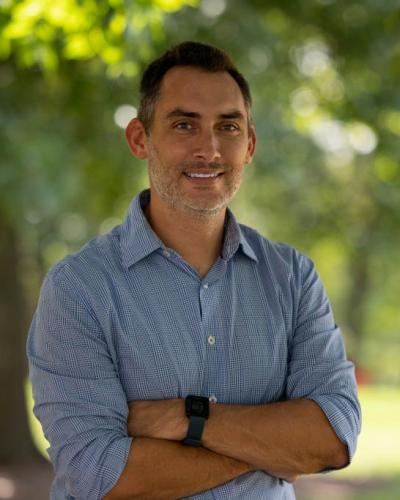 A man stands with his arms crossed, smiling, in front of a park-like setting with trees and grass.