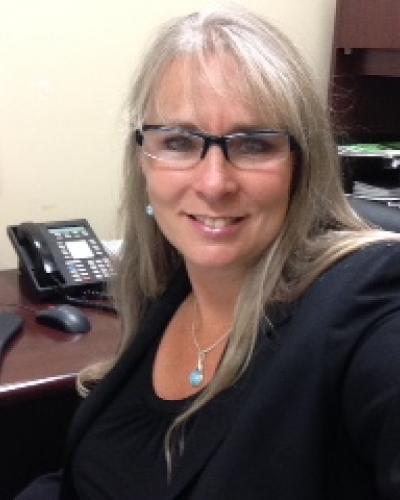 A woman in professional attire smiles while taking a selfie in an office.