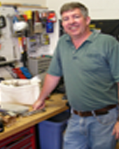 A man stands in a workshop, smiling and resting a hand on a workbench.