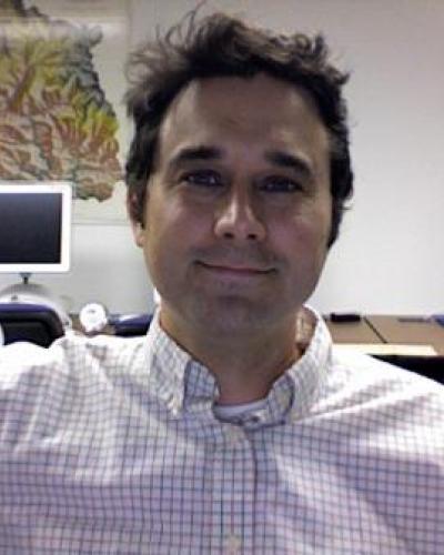 A man in a white and blue checked shirt sitting in an office with computers in the background.