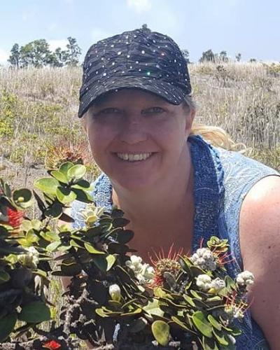 A woman wearing a hat smiles while holding plants in front of a field.