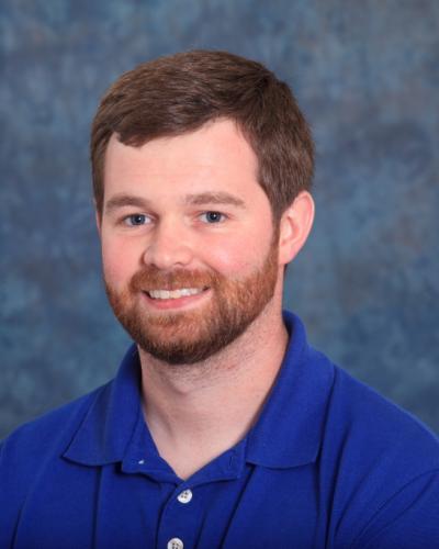 A man with brown hair and a beard wearing a blue polo shirt smiles while looking at the camera.