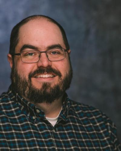 A man wearing glasses and a plaid shirt smiles while looking at the camera.