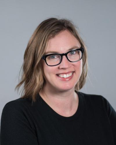 A woman with shoulder-length hair and black-framed glasses smiles while wearing a black shirt.