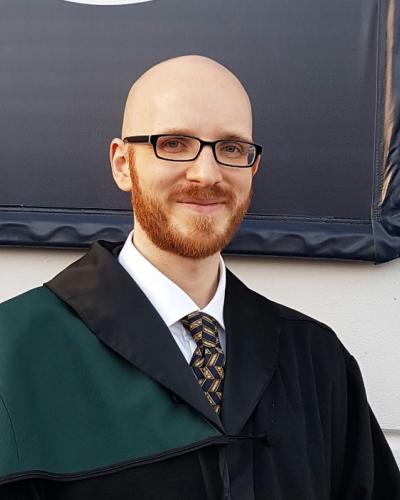 A man wearing formal academic attire smiles while posing in front of a white wall.