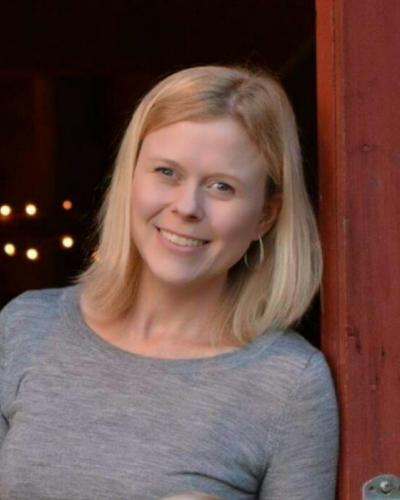 A woman with blonde hair and a gray shirt smiles while leaning against a door frame.