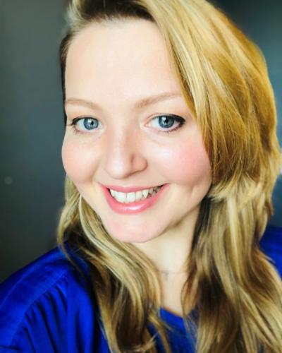 A woman with long, wavy blonde hair and a blue shirt smiles while looking at the camera.