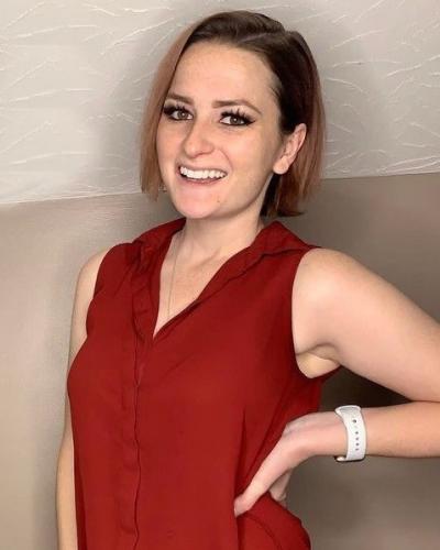 A woman smiles while posing with one hand on her hip, wearing a red sleeveless top and a white watch.