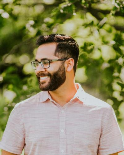 A man with short dark hair and a beard, wearing a light-colored shirt and glasses, stands in front of a green background.