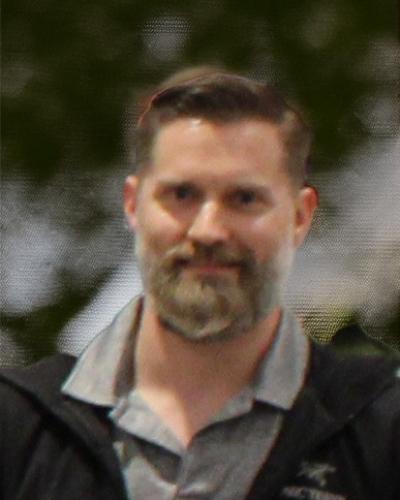 A man with short hair and a beard wearing a gray shirt and black jacket.