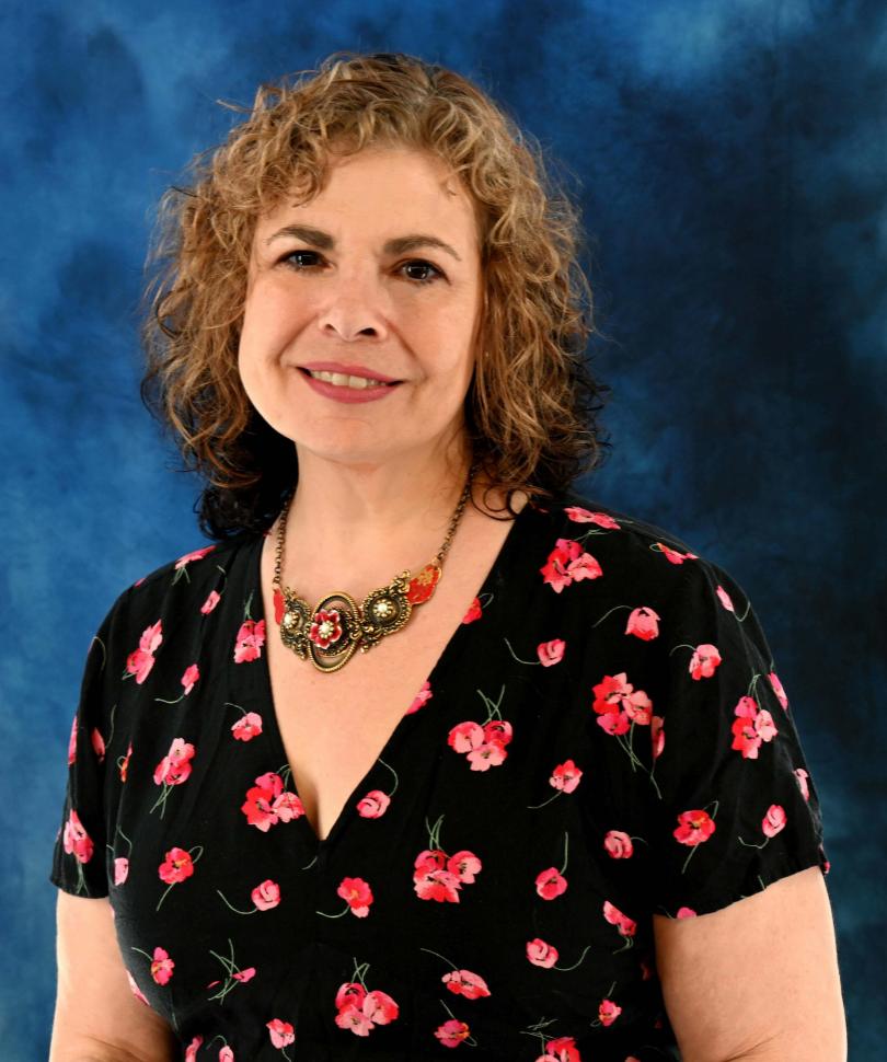 Dr. Georgia Hodes standing in front of a blue tie-dyed like background smiling. She's wearing a black dress with pink and red flowers throughout. She's also wearing an off gold necklace with red flowers.